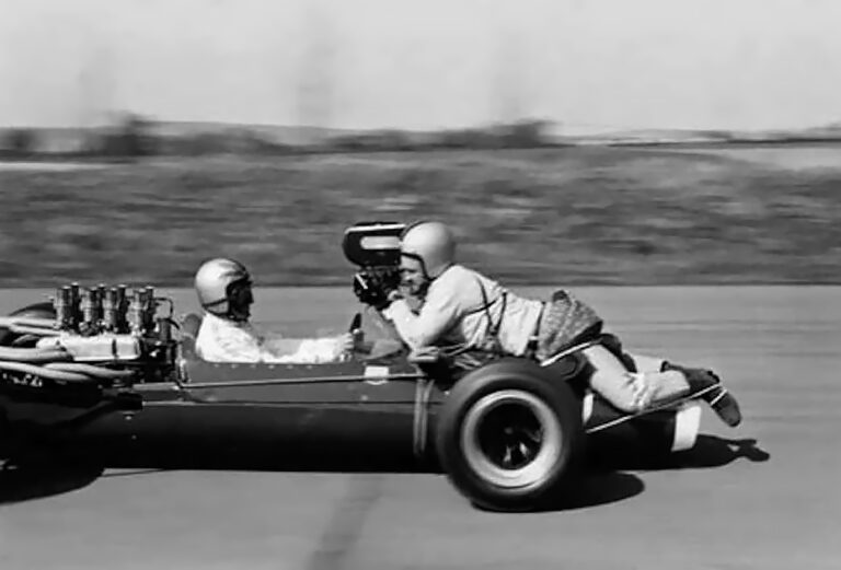An old F1 car with a camera man on the front filming the driver